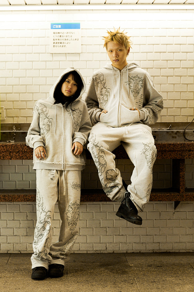 Two models in the Gray Sigil Zip Hoodie and matching pants seated against a tiled wall
