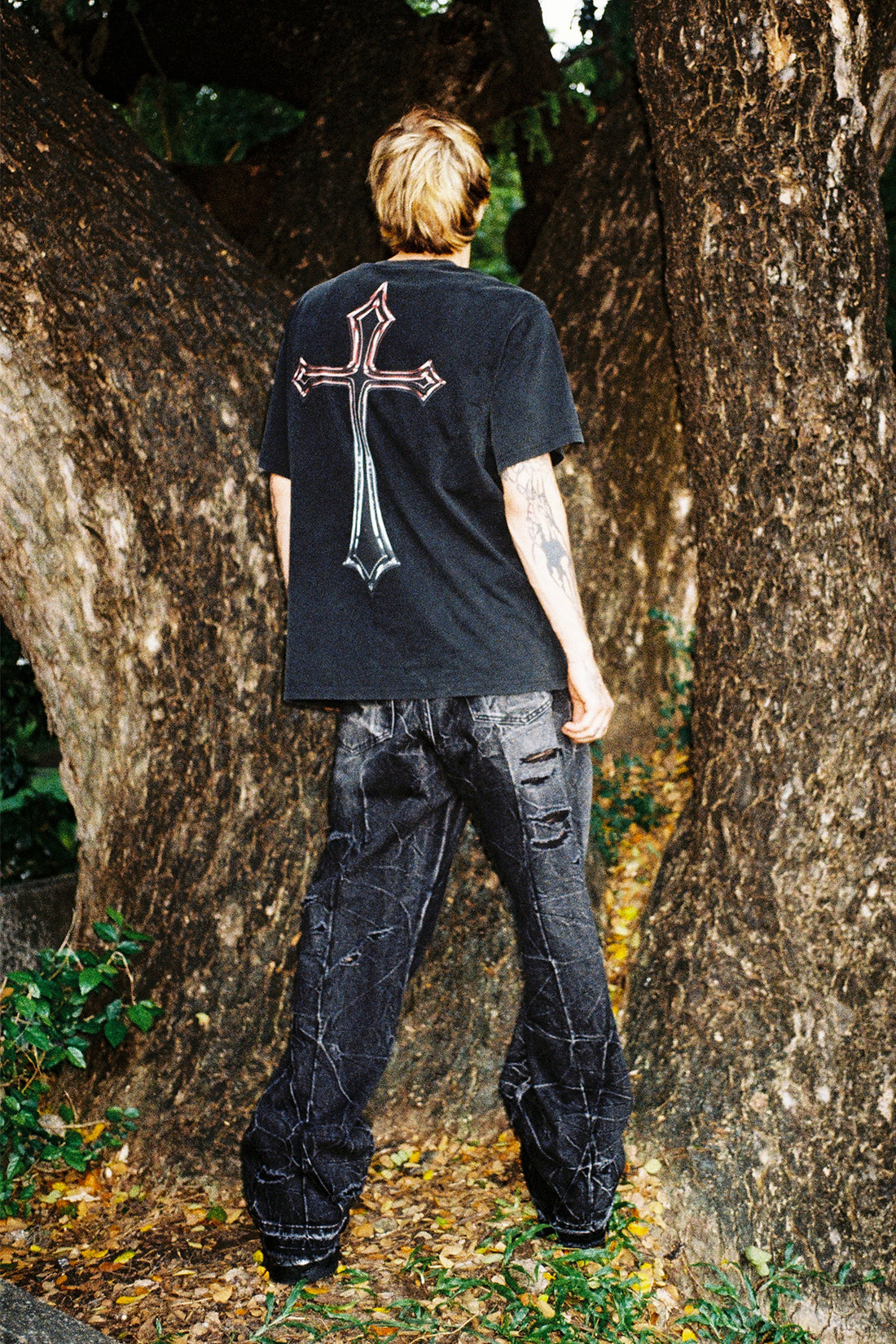 Full-length back view of the Cross Tee with distressed black jeans in a wooded setting