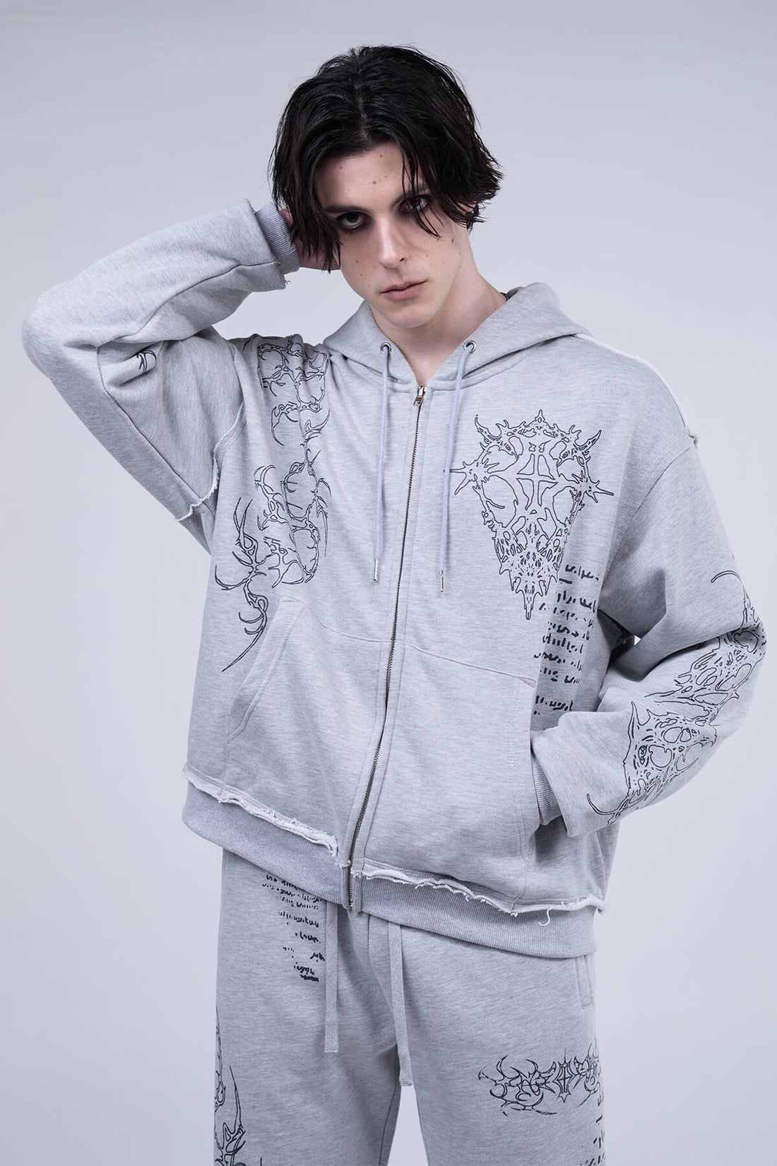 Model wearing the Gray Sigil Zip Hoodie with dark tribal embroidery, hand raised to his head