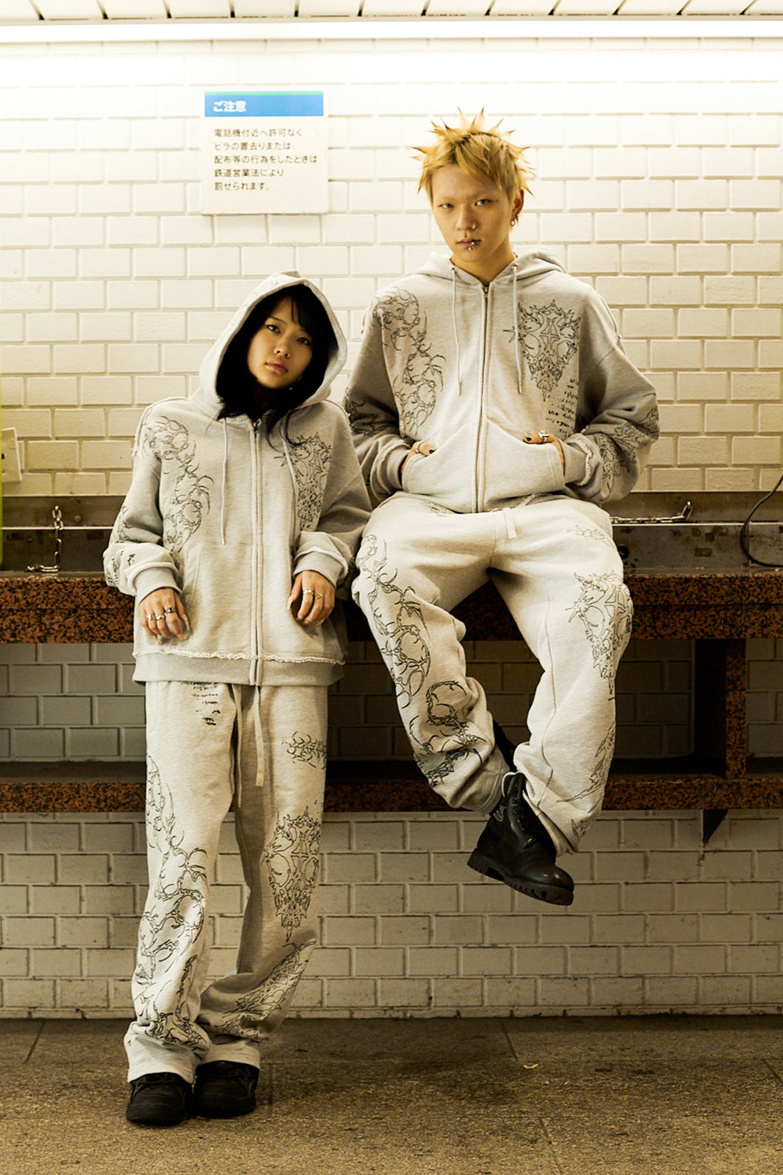 Two models in the Gray Sigil Zip Hoodie and matching pants seated against a tiled wall