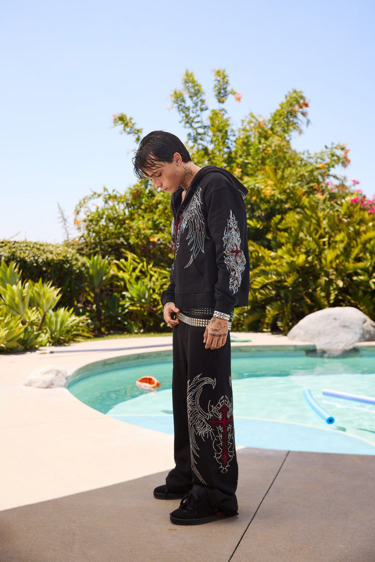 Side view of the Red Cross Zip Up Hoodie and rhinestone sweatpants outdoors by a pool